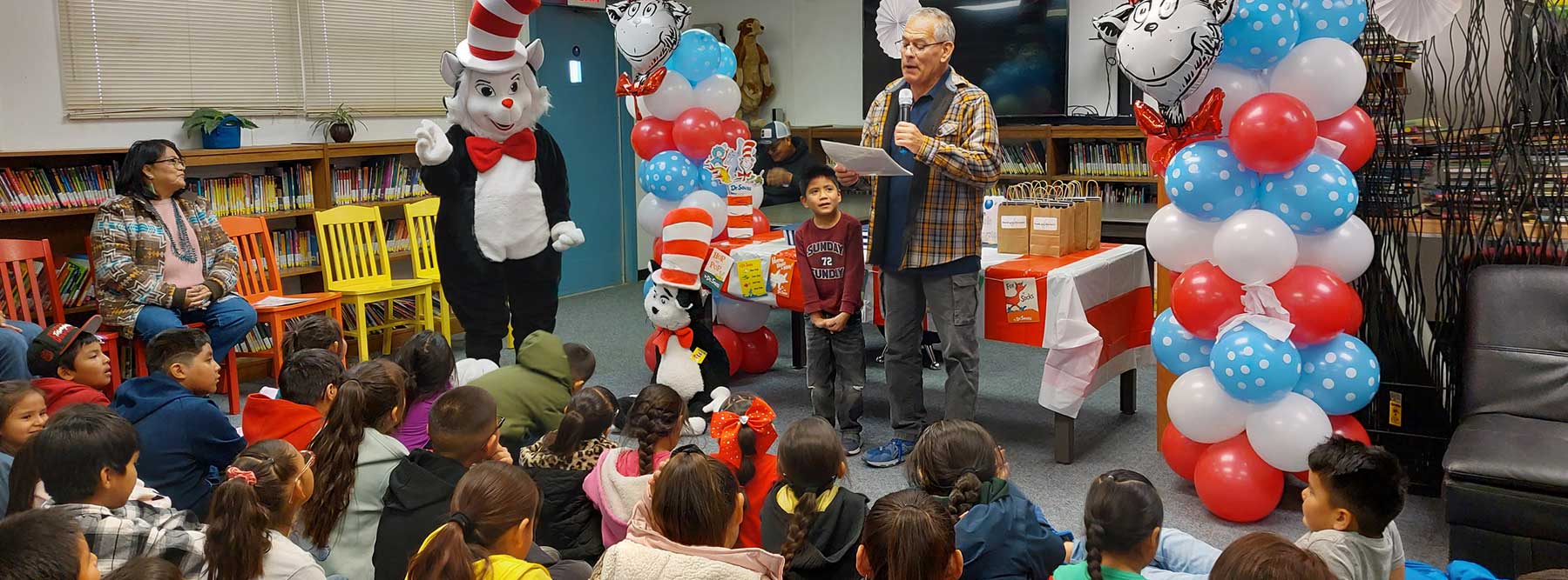 Children sit in a library listening to a speaker beside a Cat in the Hat character and colorful balloon decorations during a reading event.