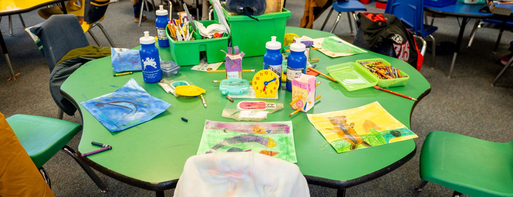A colorful classroom table with paintbrushes, crayons, and scissors on it. There are chairs around the table.