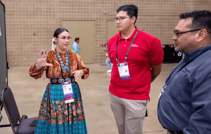 A student in a ribbon skirt and turquoise jewelry shows her research laid out on a cork board to two other people.