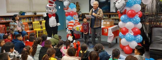Children sit in a library listening to a speaker beside a Cat in the Hat character and colorful balloon decorations during a reading event.